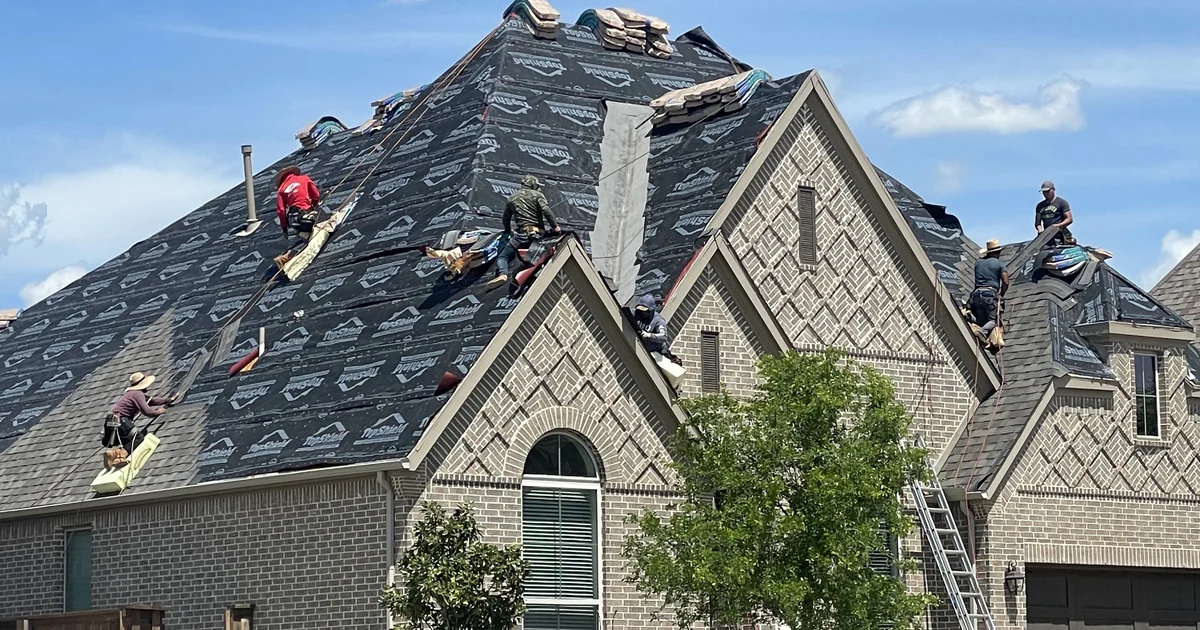 Aerial drone photograph of a North Texas custom home reroof in progress by Frame Restoration, with synthetic underlayment fully installed and architectural shingle bundles staged across the roof planes under a clear DFW sky