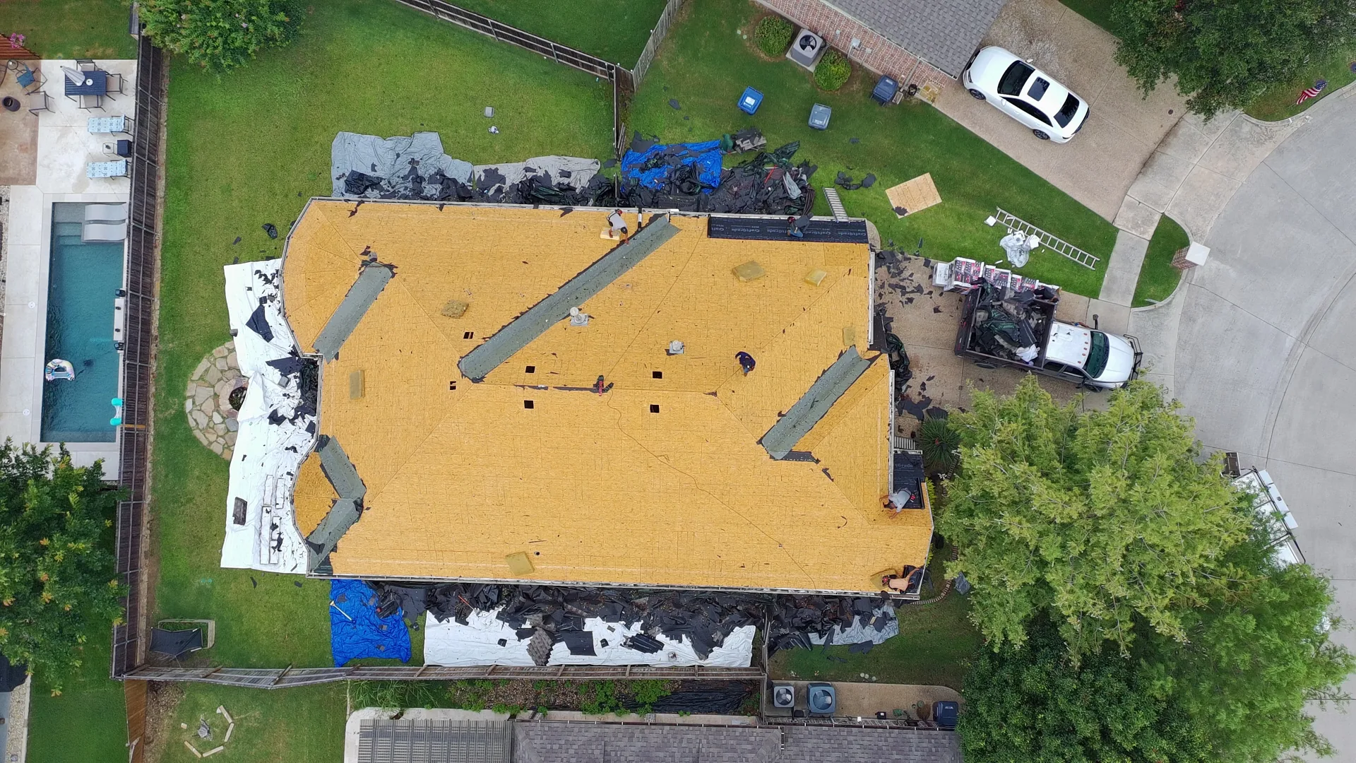 Top-down drone photo of a McKinney, Texas home with the roof torn off to bare wood decking during a Frame Restoration roof replacement, with debris staged for haul-off