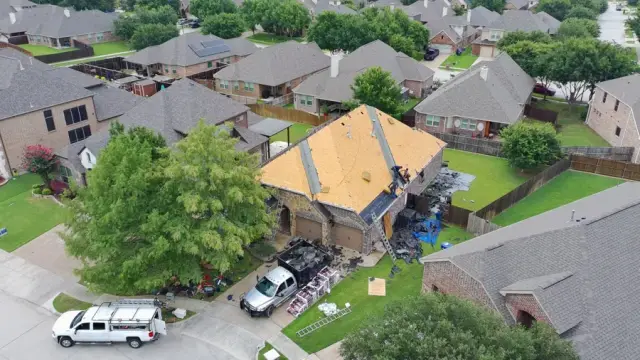 Aerial drone view of a two-story McKinney, Texas home with the roof torn off to bare wood decking during a Frame Restoration roof replacement, work van and dump truck staged in the driveway