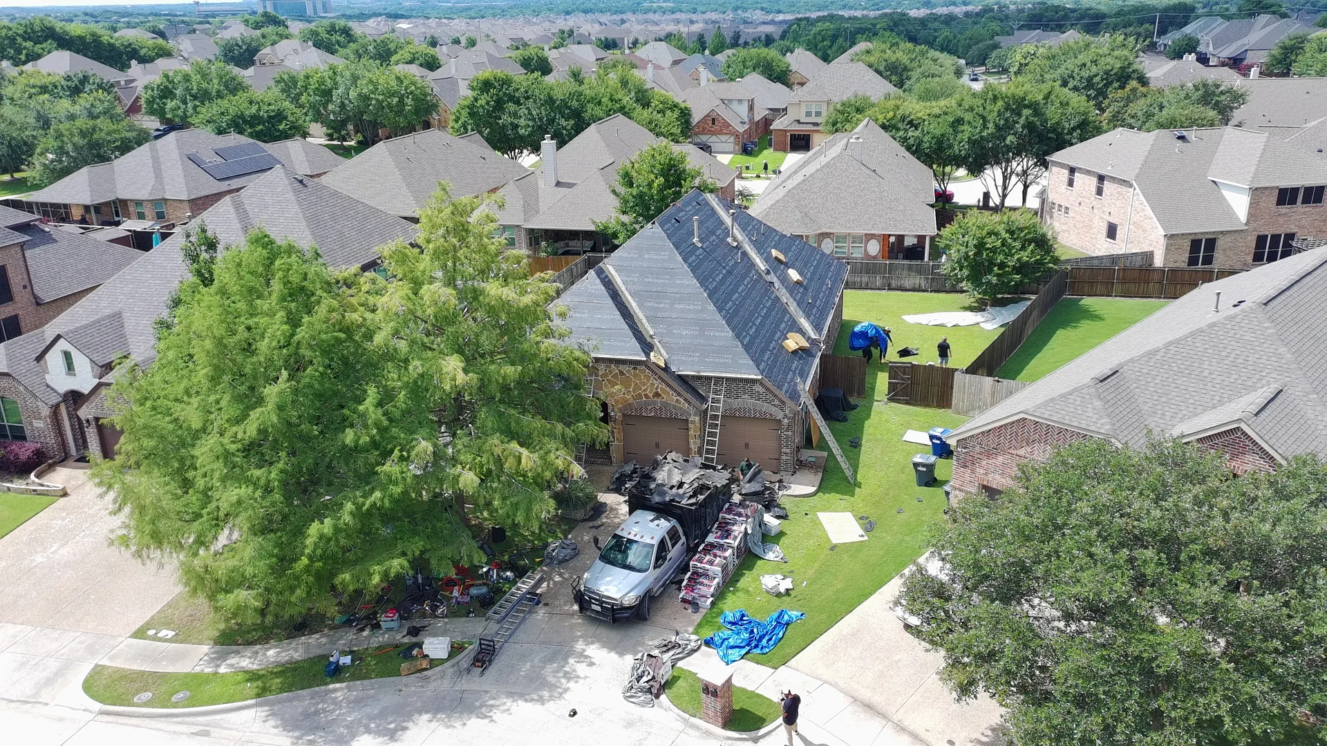 Aerial drone view of a two-story McKinney home with synthetic underlayment installed and a ladder staged, set in a North Texas neighborhood